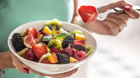 Getty Images A woman eats a colourful bowl of fruit salad. She is holding a fork, which has a strawberry on it, in her left hand.