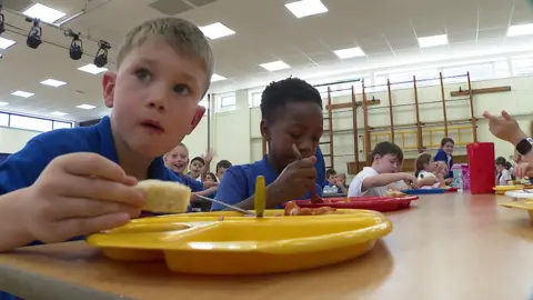 Spencer sitting on the left holds a piece of bread and looks to the side while Harry on the right puts his fork on his red plate, with other children seen waving behind them in the school hall