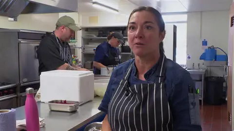 Nerissa Buckley looks emotional as she wears a blue top and striped blue and white apron as she stands in a school kitchen, with a man and woman behind her preparing food.