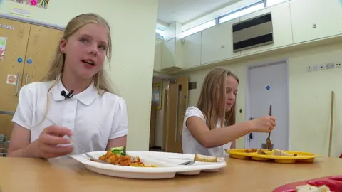 BBC Amber is dressed in a white polo top and places her fork into pasta on a white plate, looking toward the camera, while another pupils sits to her side eating from a yellow plate