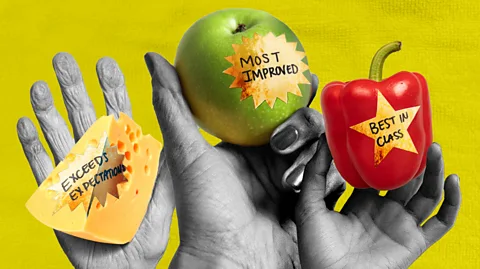 Serenity Strull/ BBC Hands holding a piece of cheese, a green apple and a red pepper with signs that read "Exceeds expectations", "Most improved" and "Best in class" (Credit: Serenity Strull/ BBC)