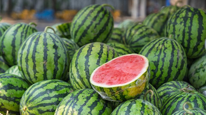 One cut watermelon half sitting on a pile of dark green whole watermelons.
