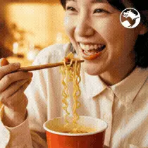Smiling woman eating noodles with chopsticks from a red cup in a warmly lit setting.