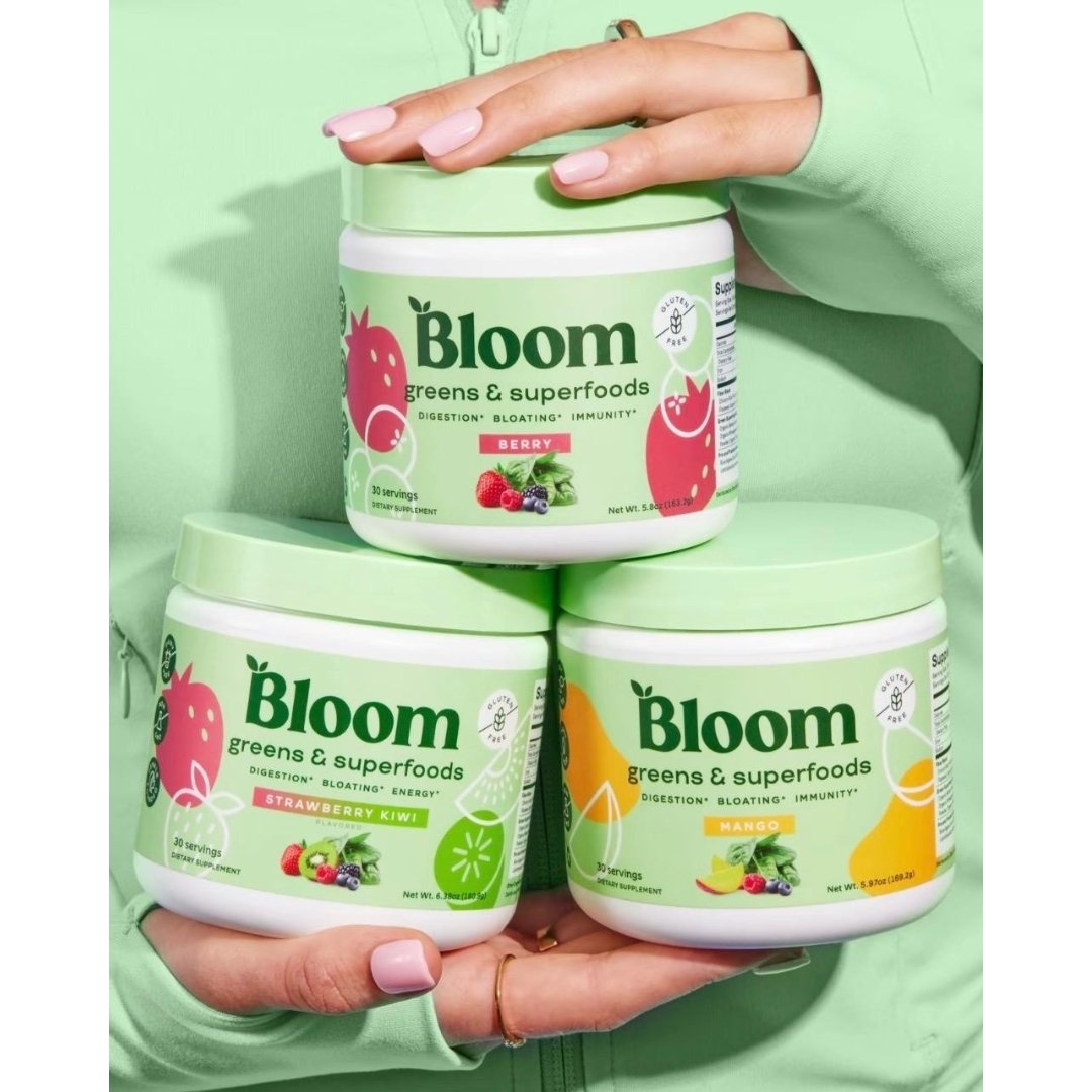 A person wearing a light green jacket holding three stacked tubs of Bloom Greens & Superfoods in Berry, Strawberry Kiwi, and Mango flavours.