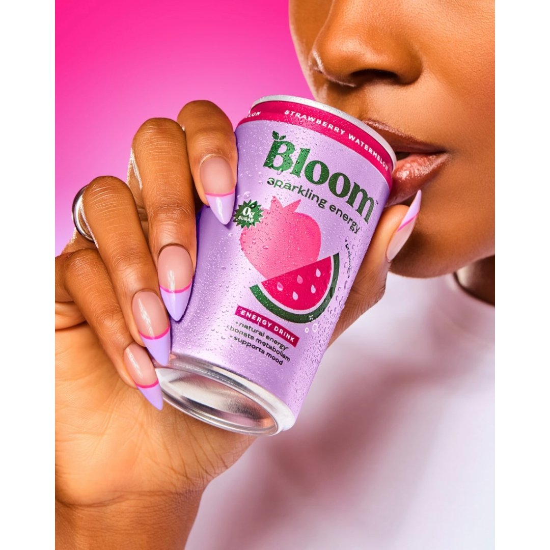 Close-up of a woman with lilac French manicure nails holding a chilled can of Bloom Sparkling Energy Strawberry Watermelon drink up to her lips against a pink background.